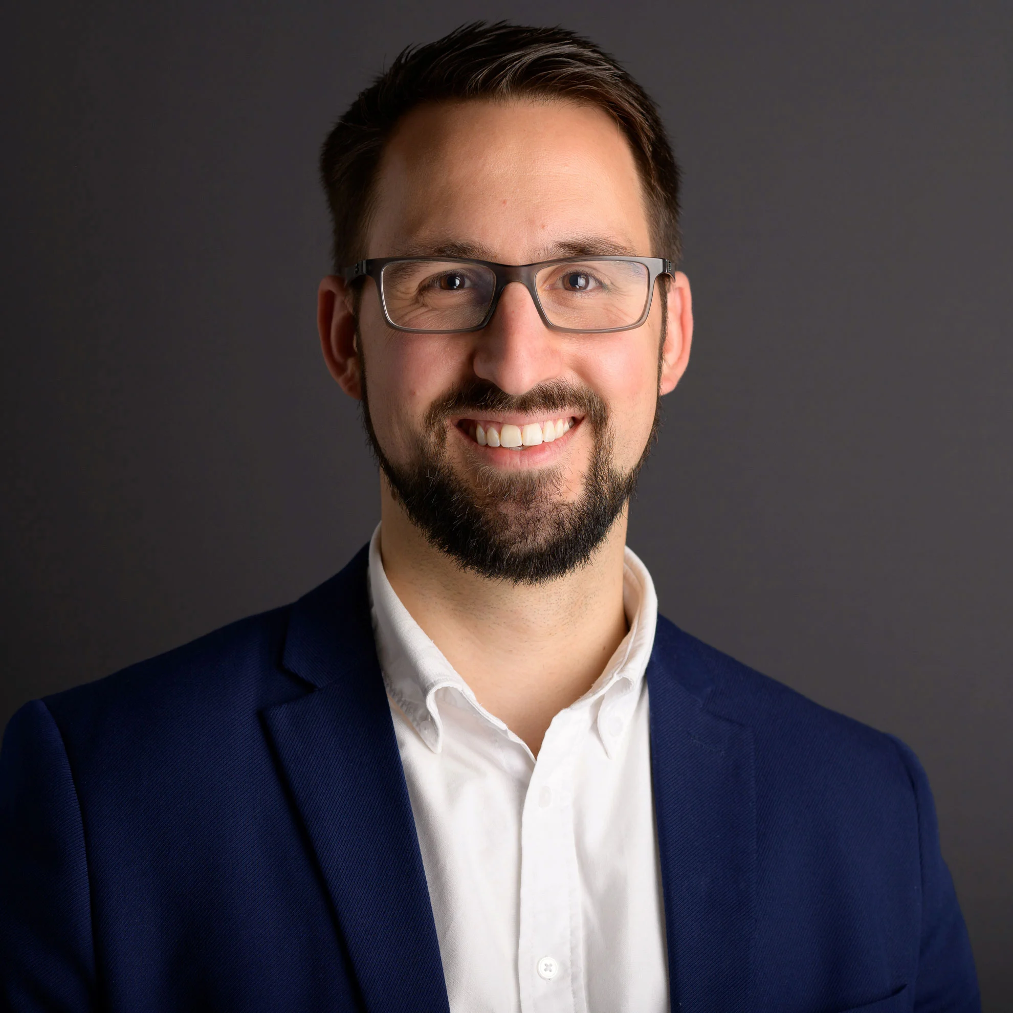 Immobilienmakler Raoul-Arthur Billan lächelt. Er trägt einen marineblauen Blazer über einem weißen Hemd und steht vor einem schlichten dunkelgrauen Hintergrund.